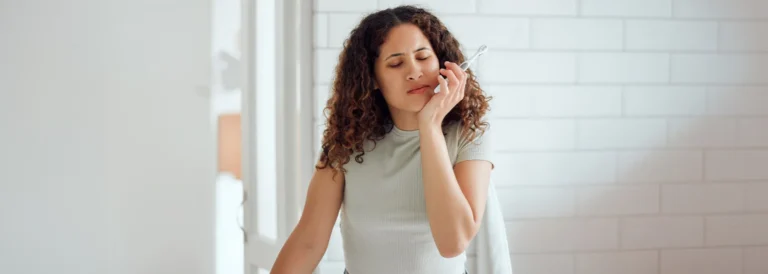 Woman holding her cheek in pain while brushing her teeth, a possible sign of a dental emergency